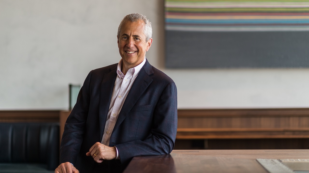 A man with gray hair and a black blazer leans against a wooden table and smiles into the camera. An abstract striped artwork is partially visible behind him.