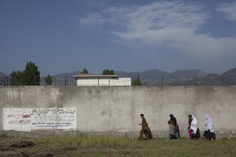 Four people walking along a long wall. A white building, mountaintops and sky are visible behind the wall.