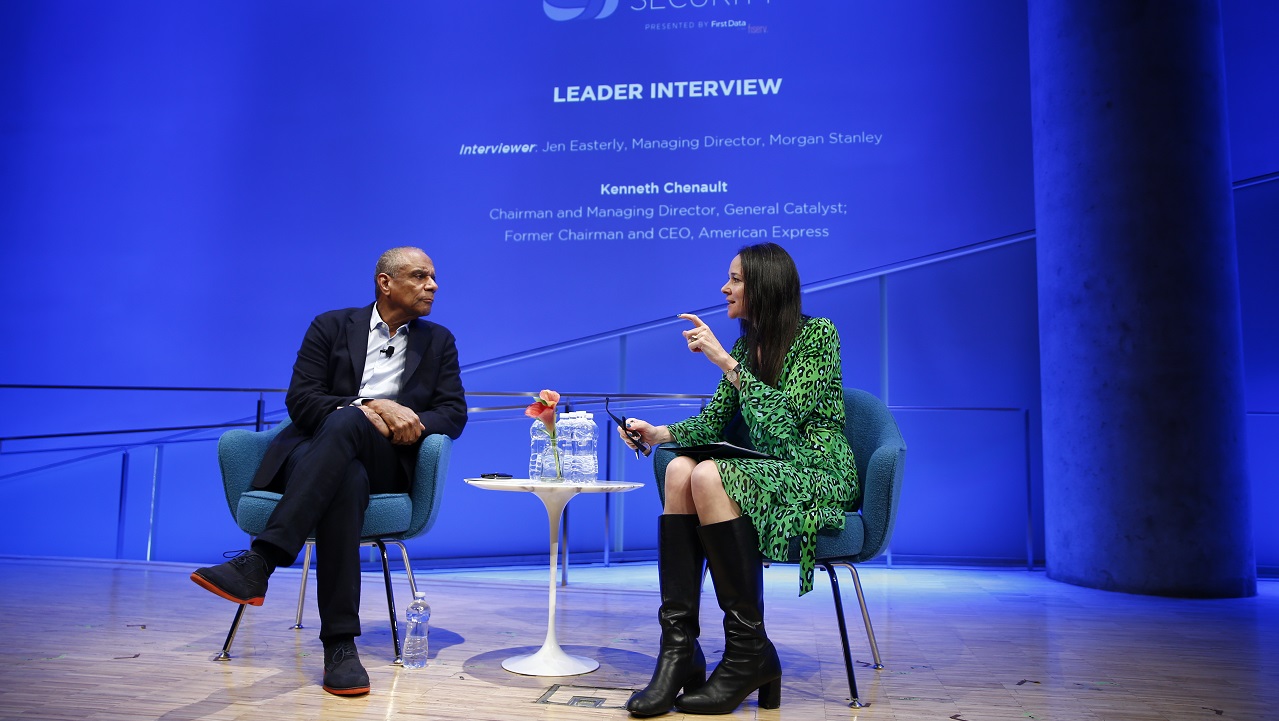 A man and a woman are seated on a stage in the auditorium of the 9/11 Memorial Museum discussing security issues before a blue display at the back of the stage.  