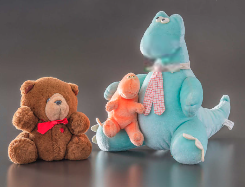 A brown stuffed bear wearing a red tie (left) and a light blue stuffed dinosaur wearing a pink tie, holding a smaller orange stuffed dinosaur