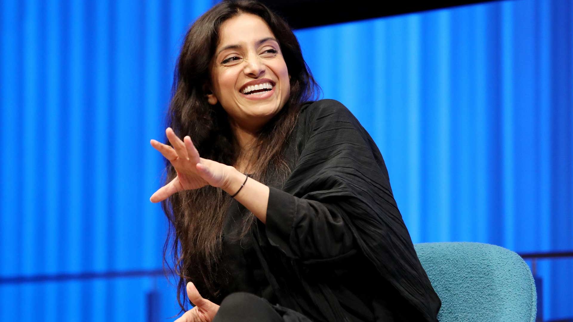 Emmy Award–winning documentarian Deeyah Khan smiles as she leans forward and gestures with her left hand onstage while speaking during a public program at the Museum Auditorium. Curtains behind her are lit up blue.