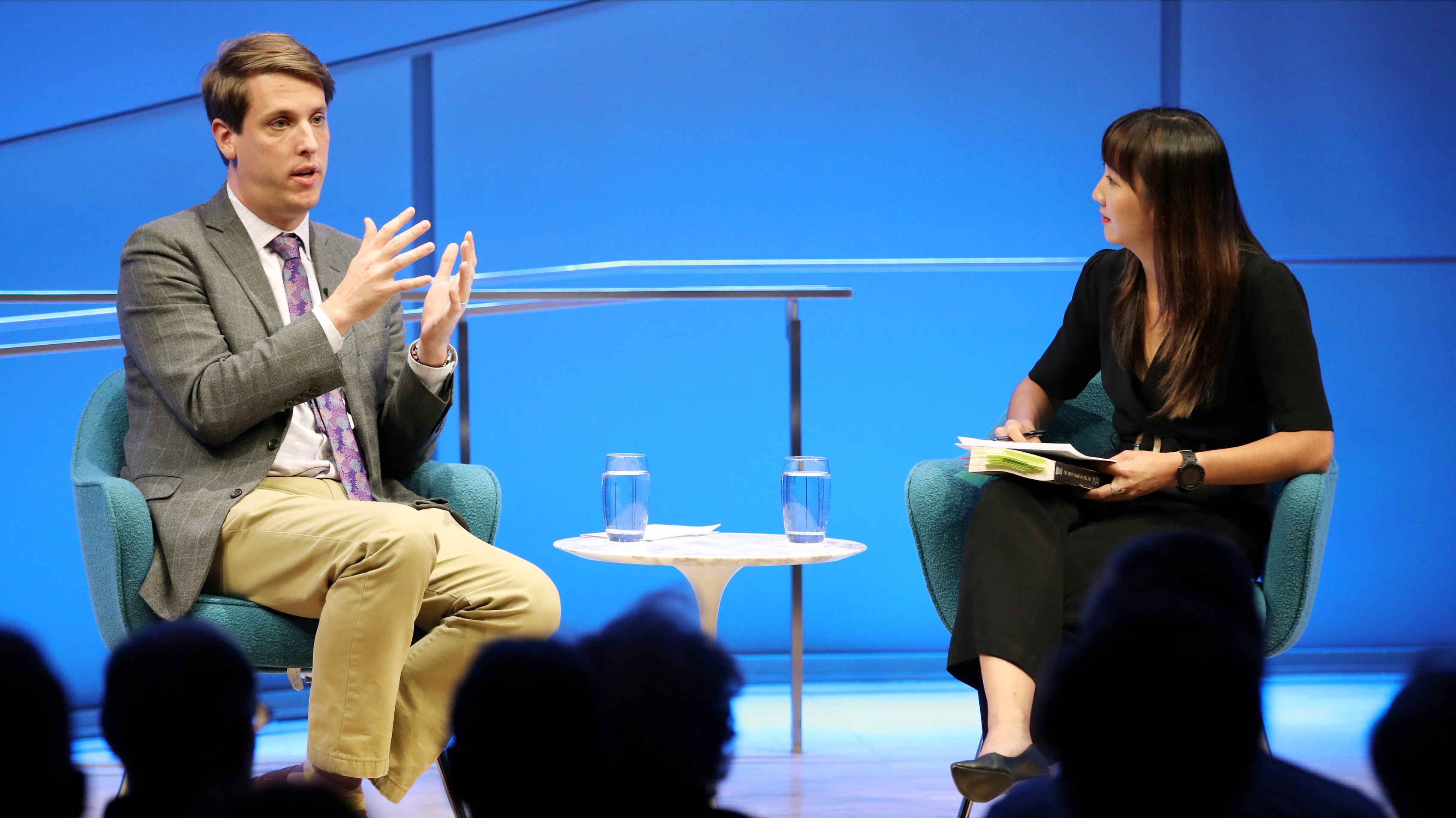 Journalist and historian Garrett Graff gestures with both hands as he speaks onstage during the public program “Only Plane in the Sky.” A woman hosting the program sits next to him as she watches on and holds a pile of papers and books. The silhouetted heads of several audience members are in the foreground.