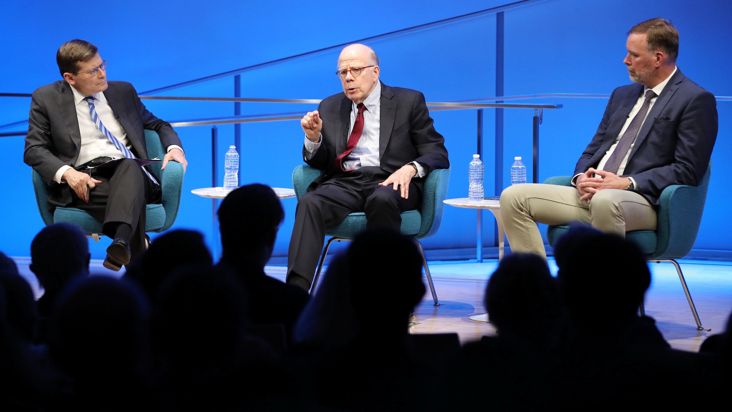 Former Acting CIA Director John McLaughlin sits between Former Acting CIA Director Michael Morell and former CIA Senior Paramilitary Officer Phil Reilly while onstage at the Museum Auditorium. McLaughlin holds up his right hand as he speaks to the crowd during the public program, “Essential Intelligence: The CIA’s Response to 9/11.” Morell and Reilly watch McLaughlin as he speaks. The silhouetted heads of audience members are visible in the foreground.