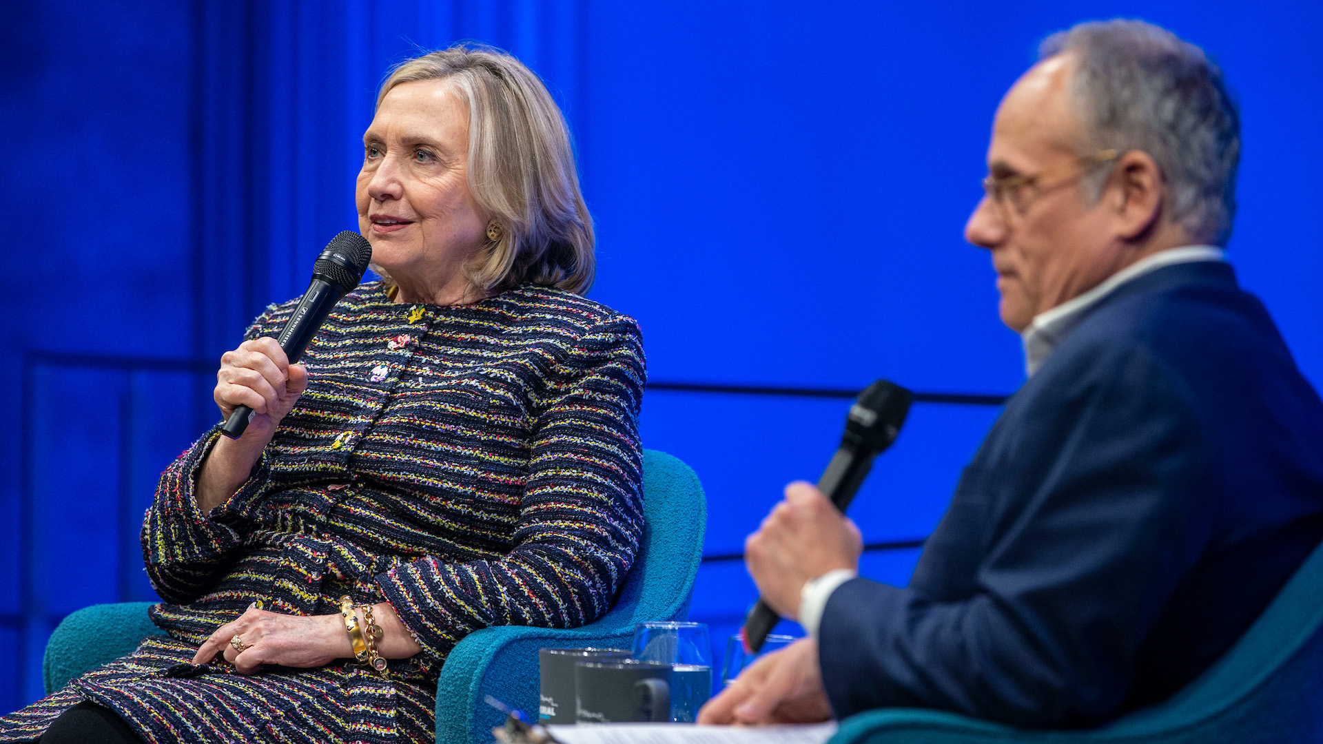 Hillary Rodham Clinton is seated, holding a microphone, to the left of a man in a navy blue suit and glasses, also with a microphone