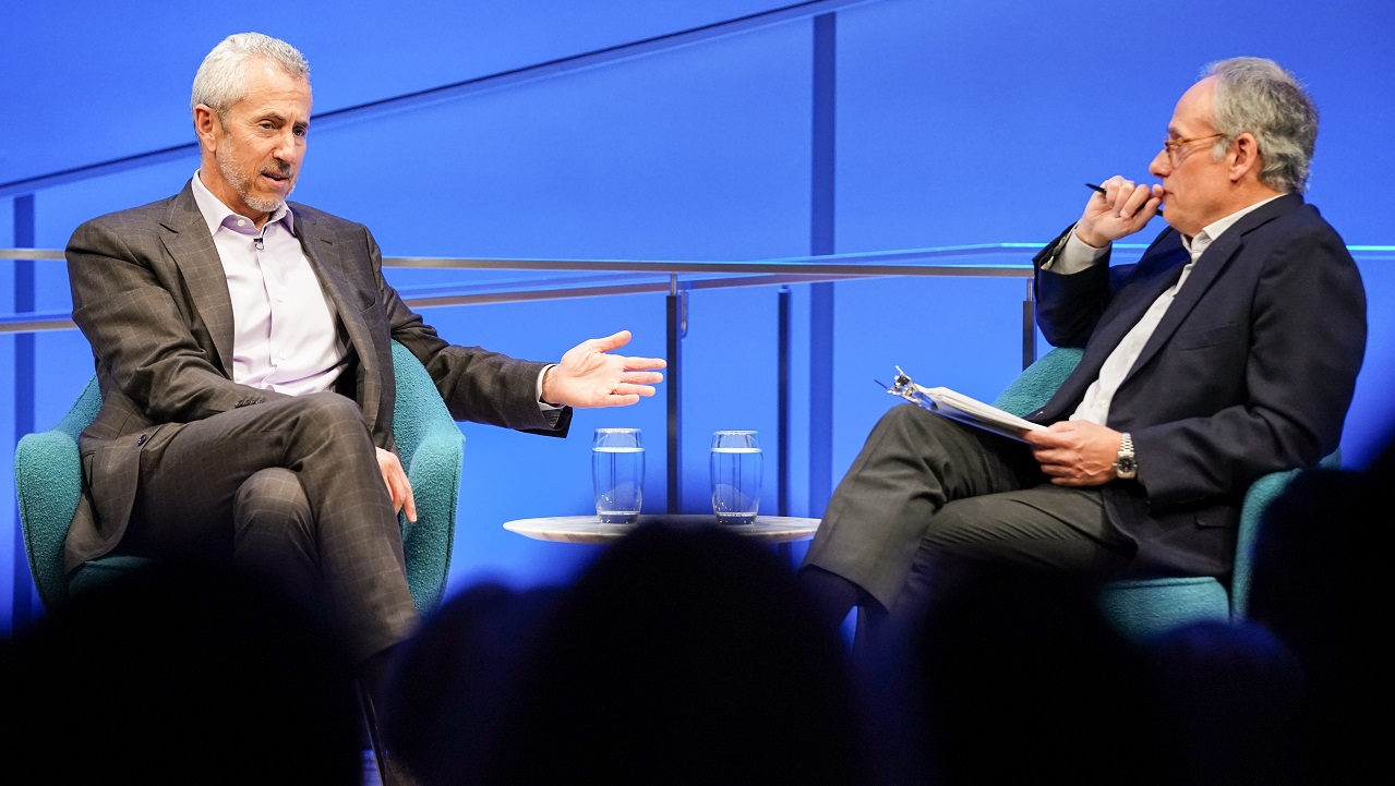 Two men in suits sit cross-legged on a blue-lit auditorium stage. One man gestures with his arms in front of him while the other looks on and listens. The heads of the audience members appear in silhouette in front of them.