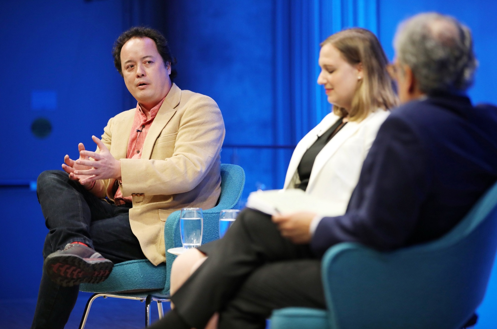 Graeme Wood of The Atlantic speaks onstage at the Museum auditorium while seated next to Devorah Margolin of George Washington University and Clifford Chanin, the executive vice president and deputy director for museum programs. Wood gestures with both hands as he looks at Chanin, who is seated in the foreground and blurred by the focus. 