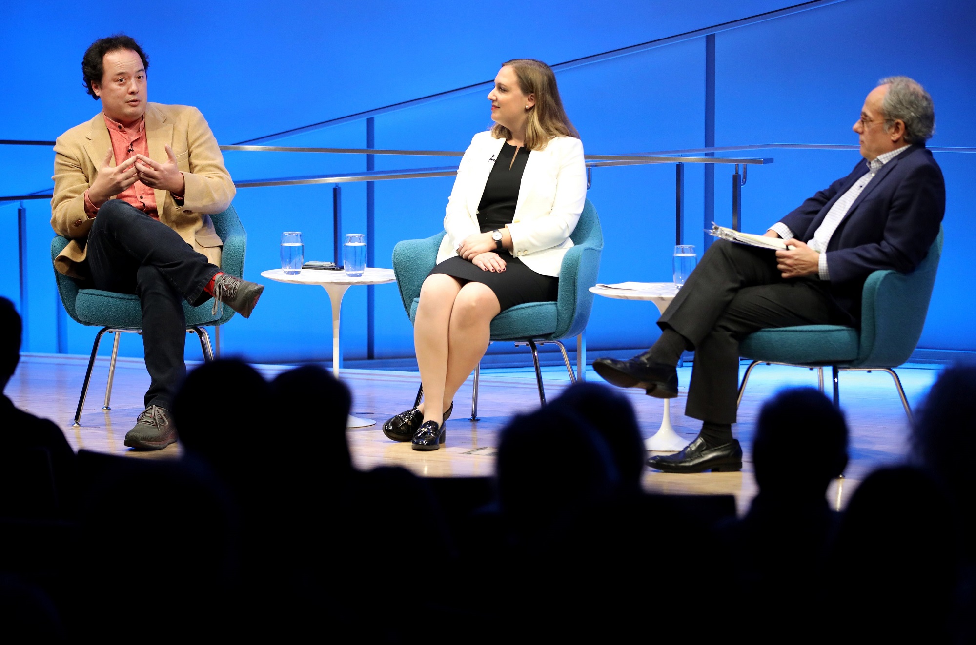 Graeme Wood of The Atlantic speaks onstage at the Museum auditorium while seated next to Devorah Margolin of George Washington University and Clifford Chanin, the executive vice president and deputy director for museum programs. The silhouettes of audience members are seen in the foreground.