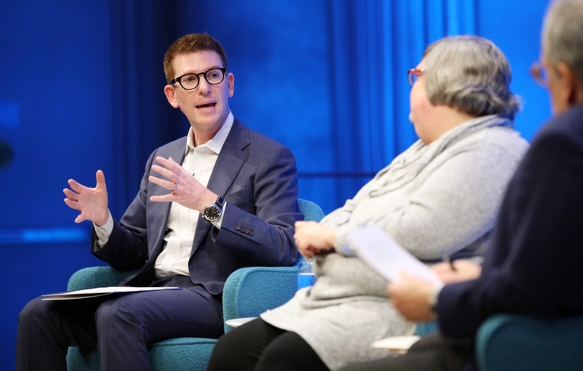 David Tessler of Facebook speaks onstage as Joan Donovan of the Harvard Kennedy School and Clifford Chanin, the executive vice president and deputy director for museum programs, watch him in the foreground. Tessler holds both of his hands up as he speaks while looking in the direction of Donovan and Chanin. 