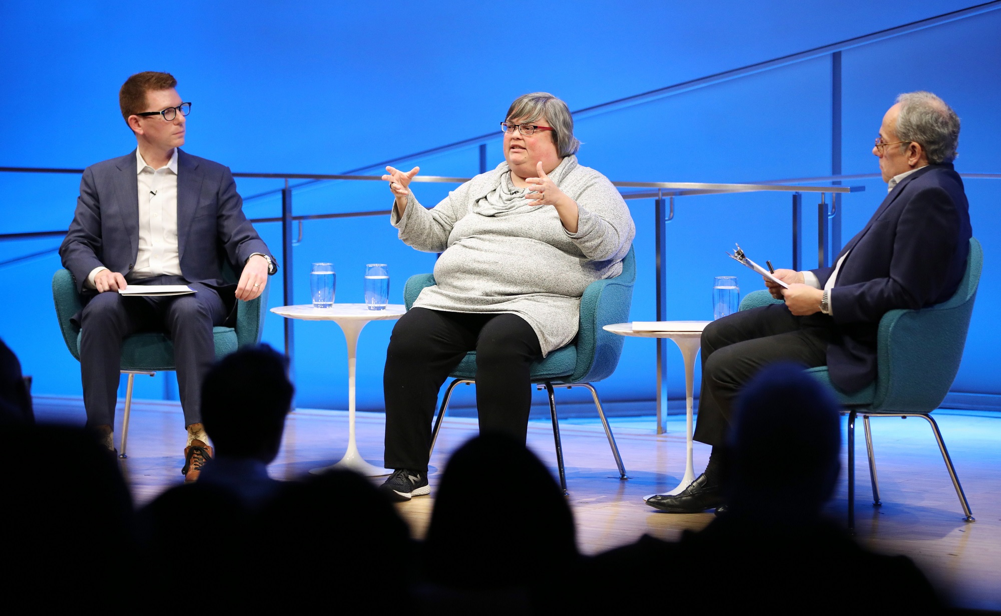 Joan Donovan of the Harvard Kennedy School gestures with both of her hands as she speaks onstage at the Museum Auditorium. David Tessler of Facebook and Clifford Chanin, the executive vice president and deputy director for museum programs, look on from her left and right. The silhouettes of audience members are visible in the foreground.