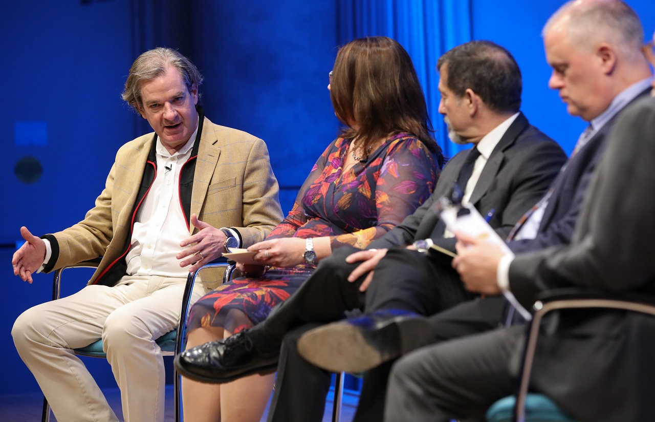 Al-Qaeda experts Peter Bergen holds out both hands and looks down in contemplation as he speaks onstage at the Museum Auditorium. Two other al-Qaeda experts, Bruce Hoffman and Mary E. Galligan, sit to his left as they listen to him speak. Mark Stout, who advised the Museum in the creation of the exhibition, is in the foreground, slightly blurred by the camera focus.