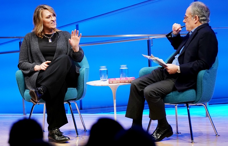 Author and former CIA analyst and targeting officer Nada Bakos sits onstage with Clifford Chanin, the executive vice president and deputy director for museum programs. Bakos puts of her left hand as she speaks to Chanin, who is holding a clipboard in his left hand and a pen with his right hand. 