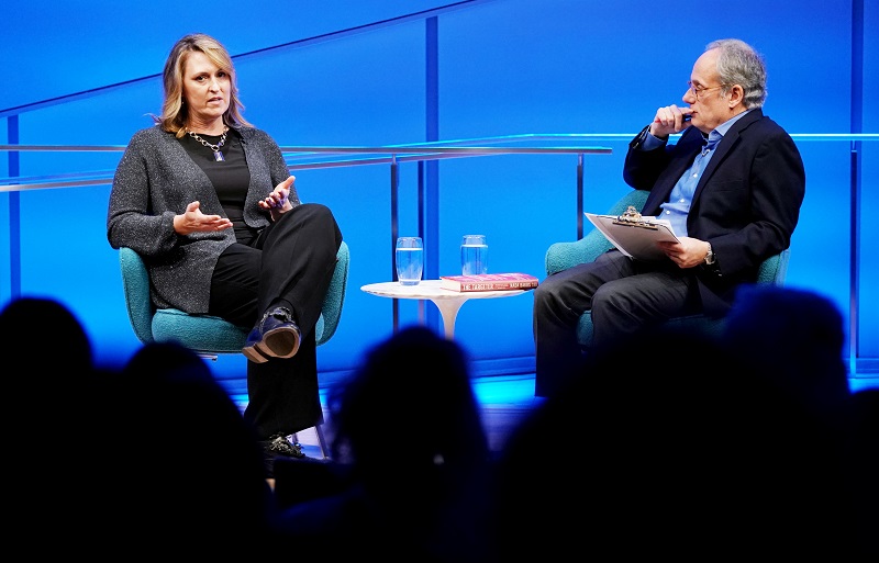 The heads of audience members are silhouetted as they look on at author and former CIA analyst and targeting officer Nada Bakos onstage at the Museum Auditorium. Bakos is gesturing with both of her hands as she speaks with her legs crossed. Clifford Chanin, the executive vice president and deputy director for museum programs, looks on from her left as he holds a clipboard and holds his right hand to his face.