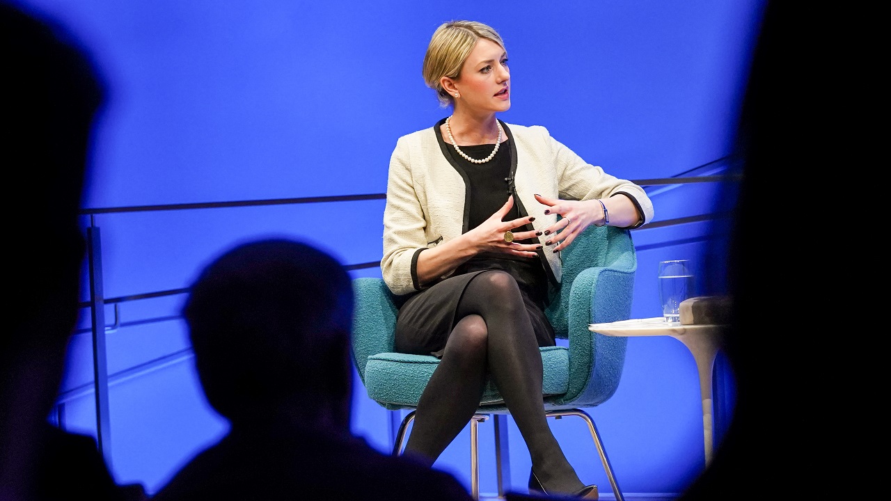 In this view from the audience, author Joana Cook can be seen speaking onstage as she sits in a chair with her legs crossed. She is holding both of her hands out as she explains something. The silhouetted heads of several audience members are in the foreground. The wall behind Cook is lit blue from the stage lights.