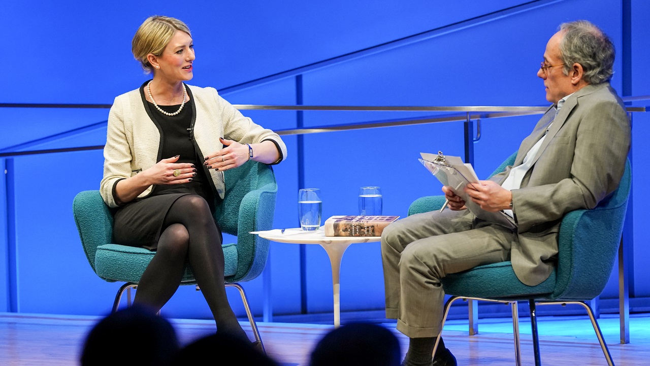 Author Joana Cook speaks with moderator Clifford Chanin onstage at the Museum Auditorium during a public program. The two of them can be seen sitting on stage together in this view from the audience. Cook is holding her hands up as she speaks with Chanin, who is holding a clipboard with both hands. The silhouetted heads of several audience members are in the foreground.