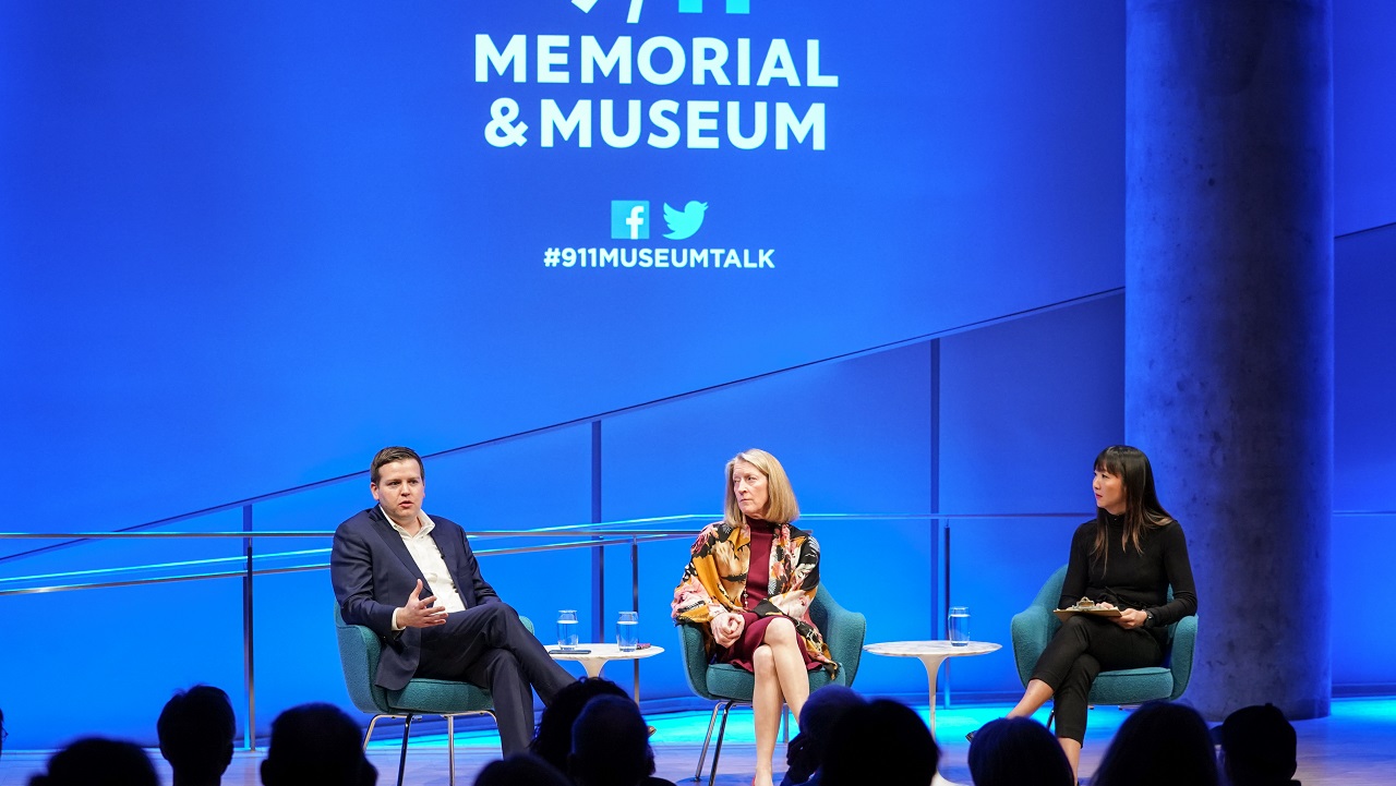 In this wide-angle view of the Museum Auditorium stage, Seamus Hughes of George Washington University’s Program on Extremism speaks as silhouetted audience members watch in the foreground. Mary McCord of the Institute for Constitutional Advocacy and Protection looks at Hughes as she sits to his left. A female moderator also sits to Hughes’ left. The 9/11 Memorial & Museum logo is projected on a wall above the three of them.