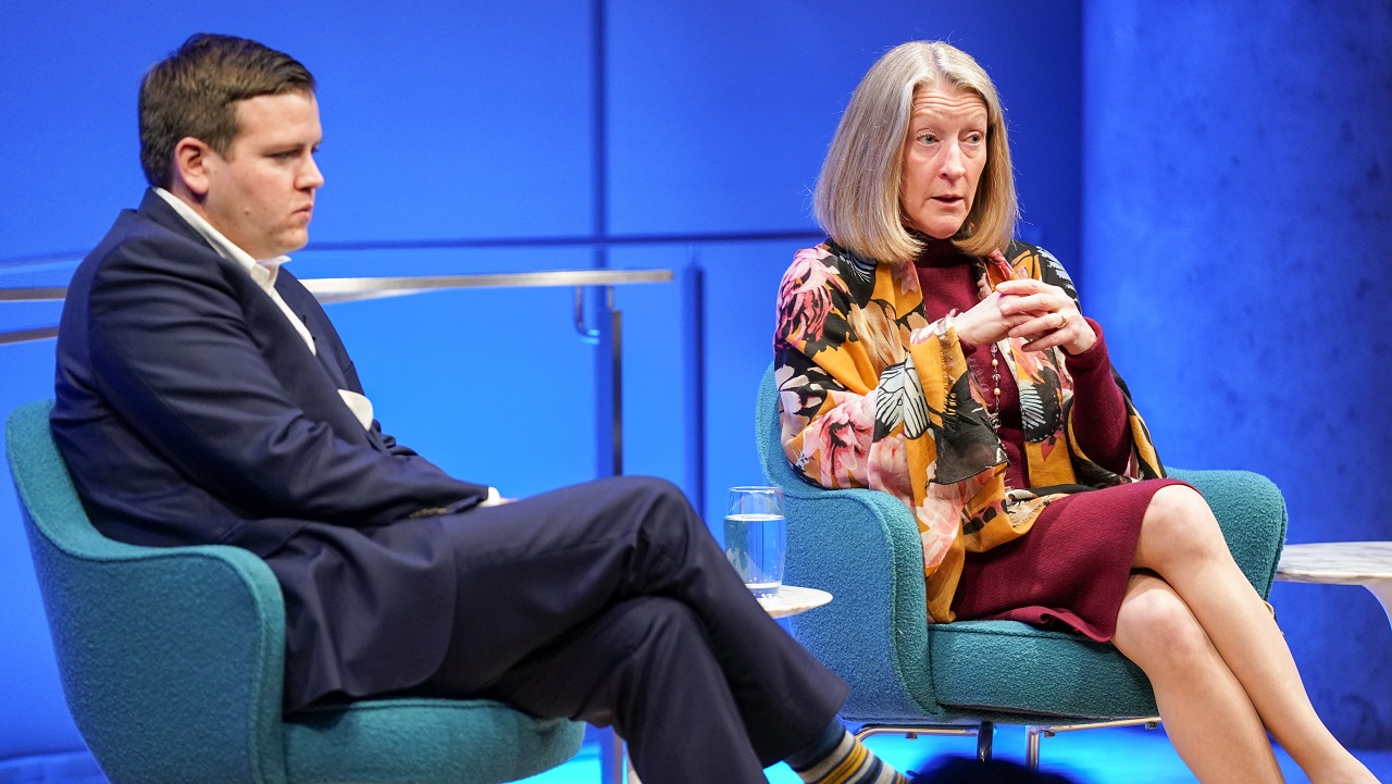 Mary McCord of the Institute for Constitutional Advocacy and Protection appears to be listening to someone out of view as she sits with her hands folded and legs crossed on the Museum Auditorium stage. Seamus Hughes of George Washington University’s Program on Extremism sits and listens beside her with his legs crossed. The wall is lit up blue behind the two of them.