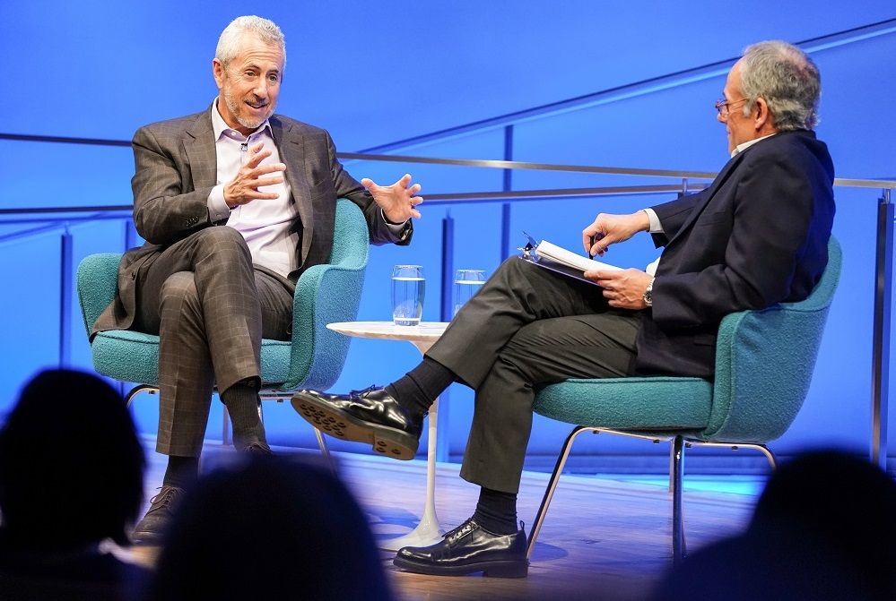 Two men in suits sit cross-legged on a blue-lit auditorium stage. One man gestures with his arms in front of him while the other looks on and listens. The heads of the audience members appear in silhouette in front of them.