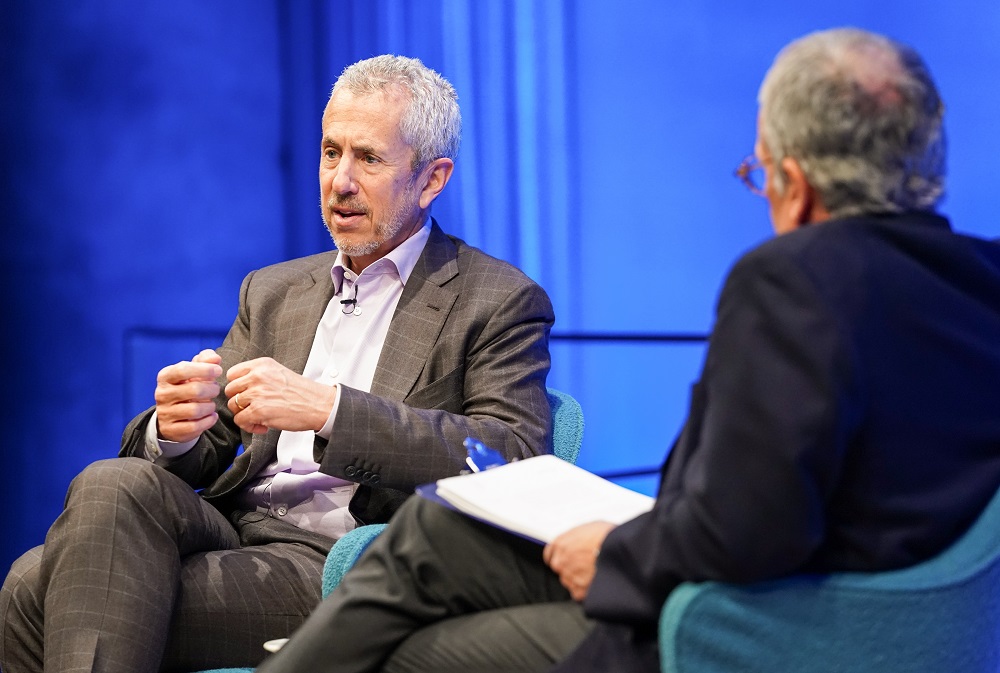 Two men in suits sit cross-legged on a blue-lit auditorium stage in this close-cropped photograph. One man's back is to the camera.