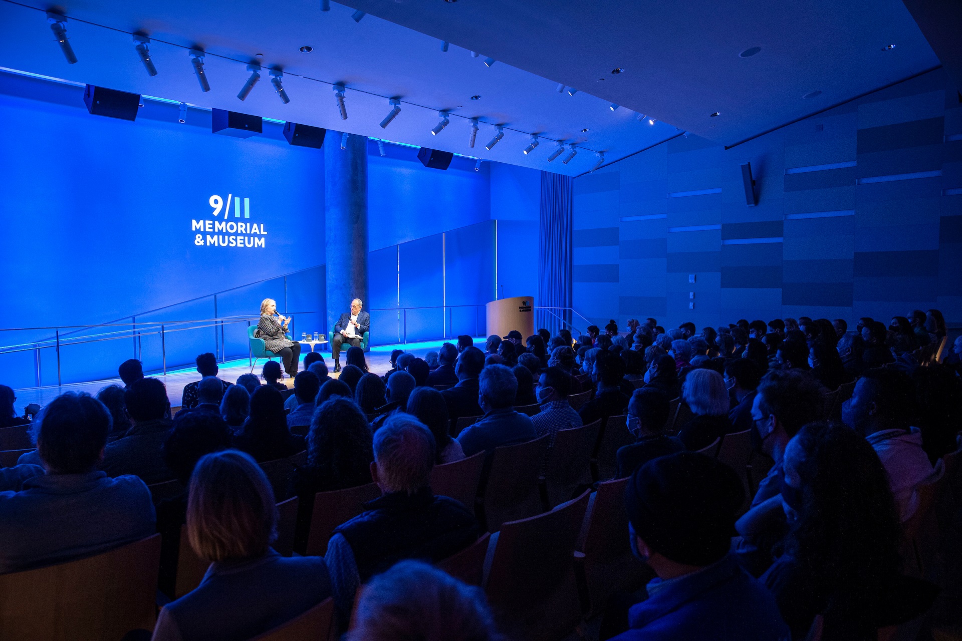 Speakers on an illuminated stage with bright blue curtain face the silhouettes of audience members