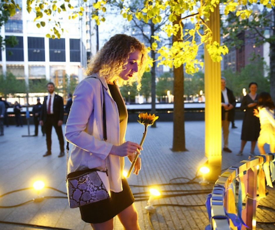 A blonde woman stands at the base of the Survivor Tree looking at tributes