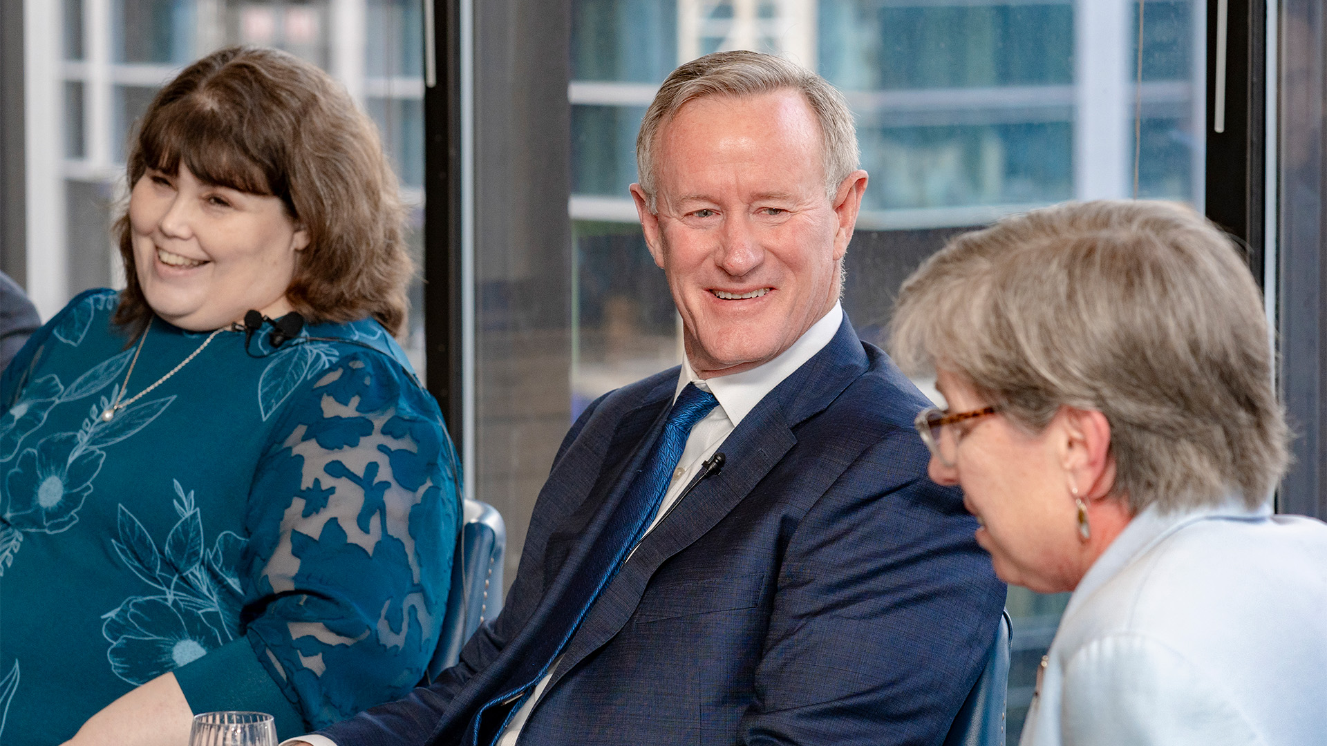 Three speakers, seen from the chest up, smiling as they discuss in front of a view of the city behind them