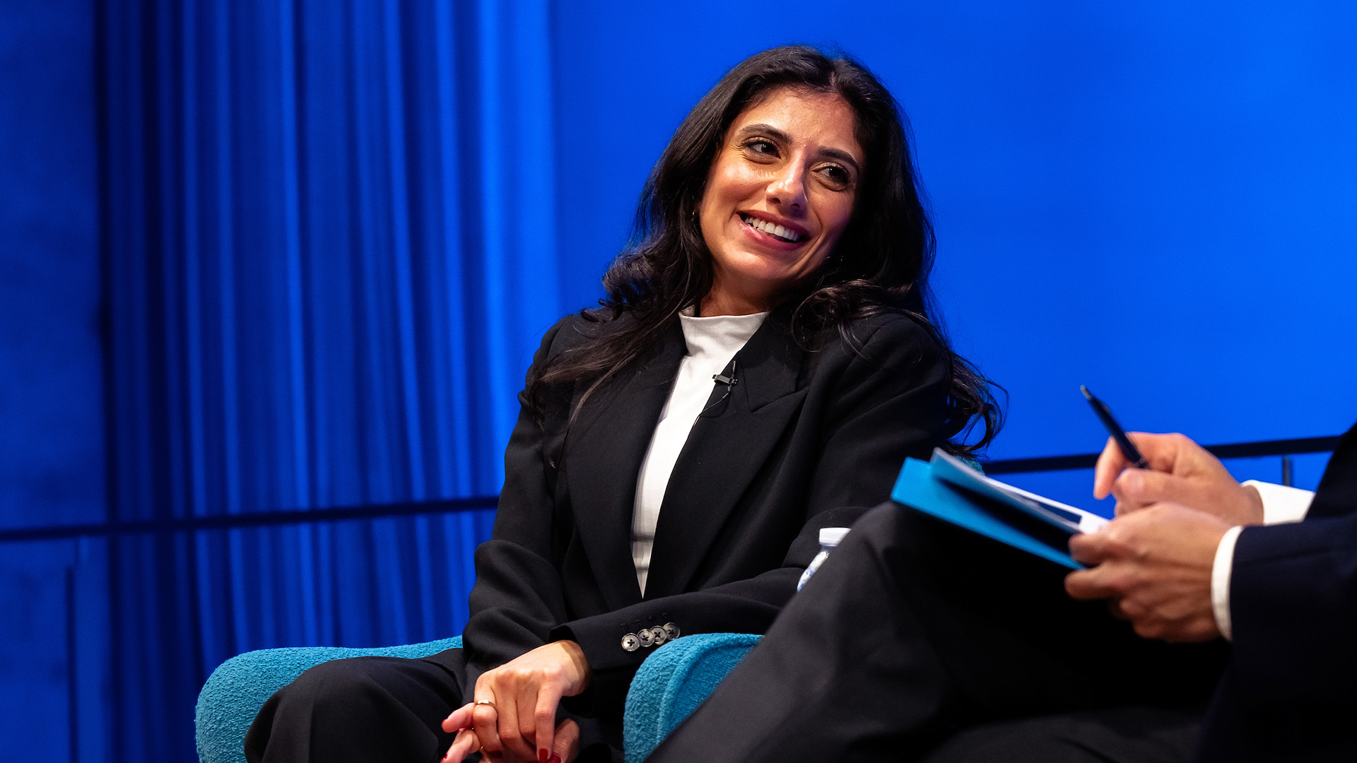 A woman with long, dark hair sits on stage, smiling, with blue curtain in background.