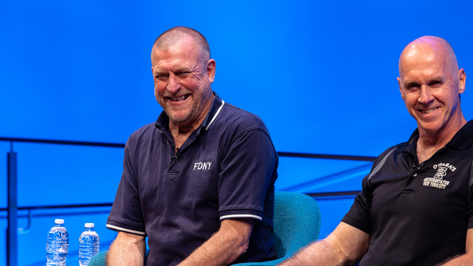 two speakers sitting in front of a blue backdrop, smiling brightly.