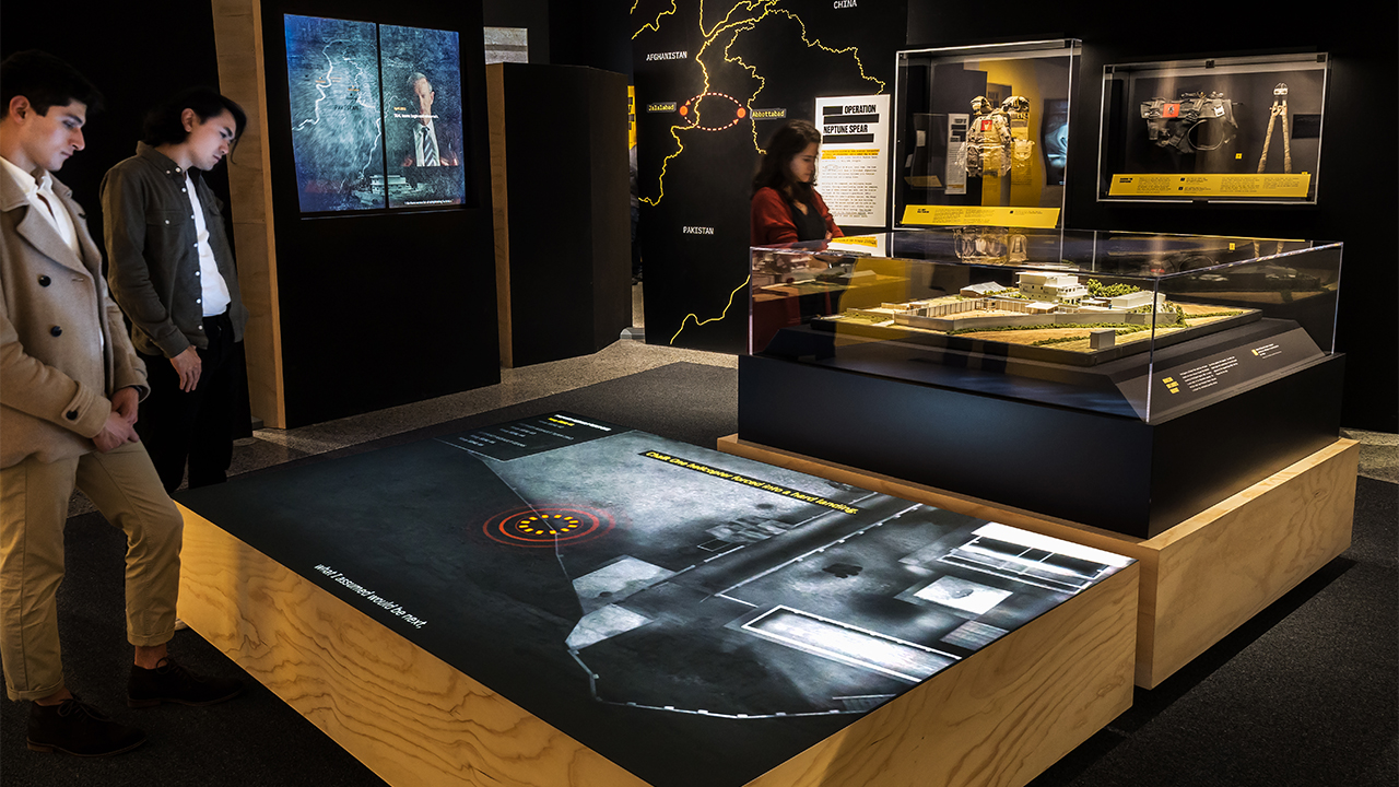 In a gallery space, two visitors watch a projected film on horizontal surface and a visitor looks at a display case. 