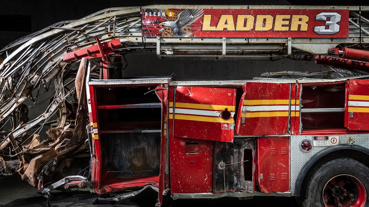 The heavily damaged firetruck of Ladder Company 3 sits in the Museum. This close-up view shows the bright red vehicle’s twisted ladder and broken compartment doors. 