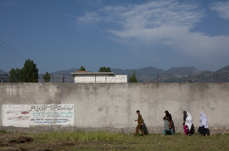 Four people walking along a long wall. A white building, mountaintops and sky are visible behind the wall. 