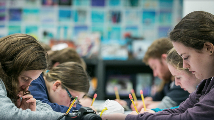 A group of students sit around a table, holding pencils, and peering down as they work. They are deeply engaged in a learning activity. 