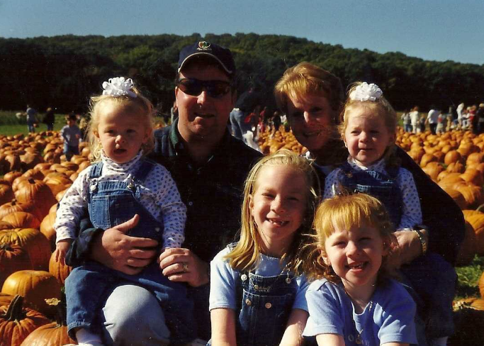 A husband and wife with four toddler and elementary-aged children sit in a pumpkin patch on a sunny day. 