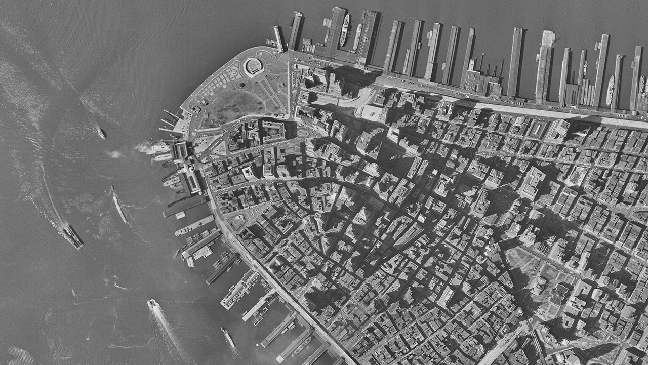 Black and white photograph with bird's eye view of lower Manhattan with piers and boats along the coast. 