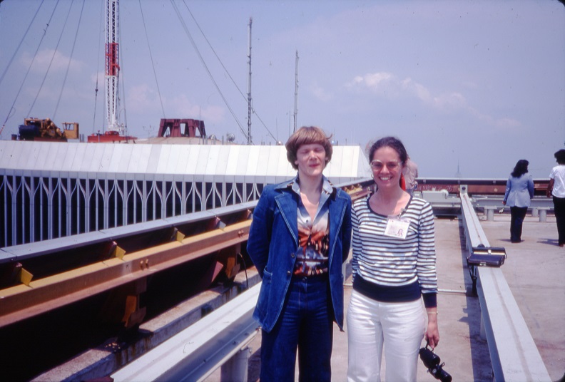 Two smiling individuals pose for the camera on a rooftop with the top part of the North Tower visible in the background. 