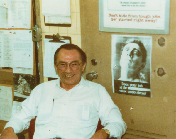 This historical photo shows Robert Kirkpatrick in a white collared shirt in his office at the World Trade Center. Papers and images are tacked onto the wall behind him.  