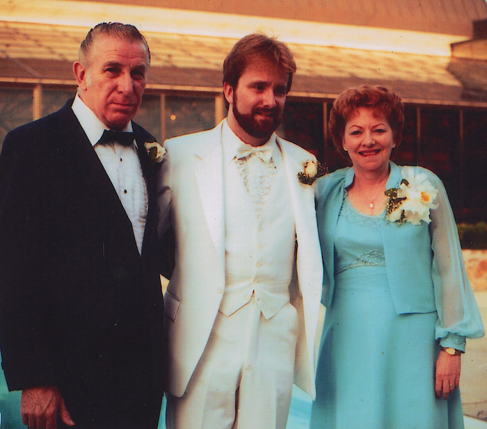 A bearded man in a white tuxedo stands between his father, on the left, in a black and white tuxedo, and his mother, on the right, in a turquoise dress 