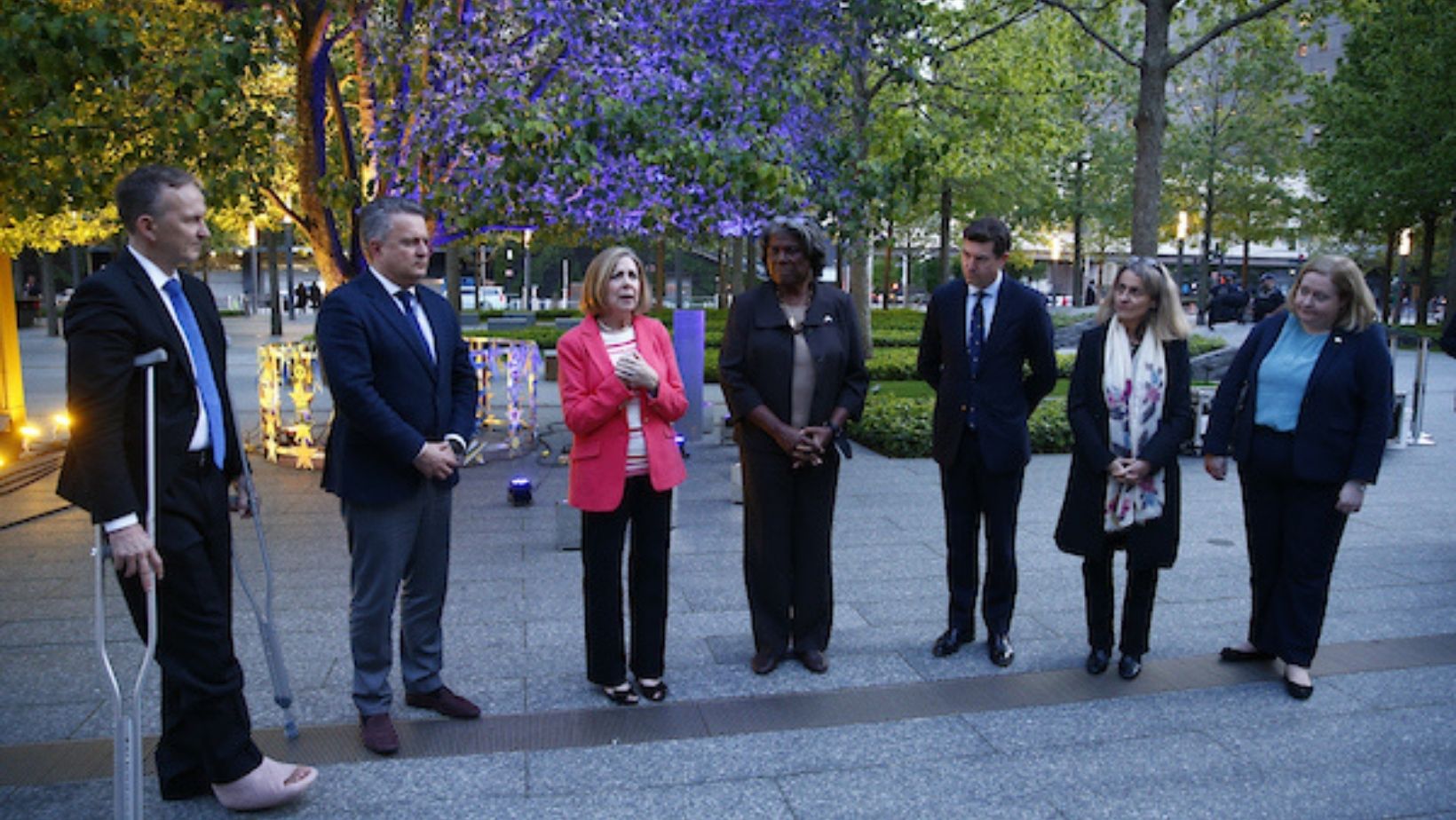 A woman in a red jacket and dark pants speaks to a group of diplomats in dark clothes 