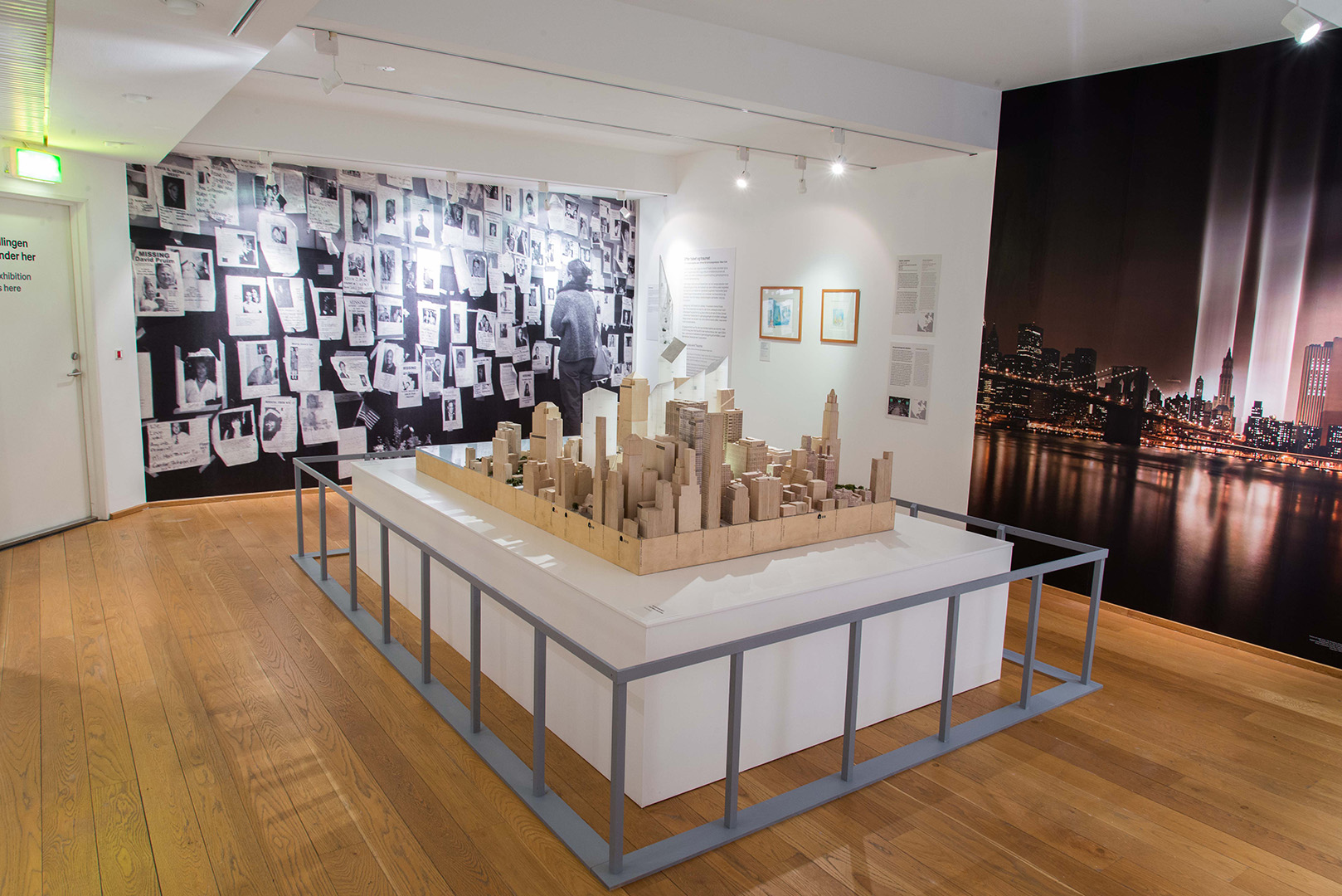 A large wooden model of downtown Manhattan in a museum gallery. On the walls behind it are a mural photo of missing persons posters and a mural photo of two columns of light beaming where the Twin Towers used to stand. 