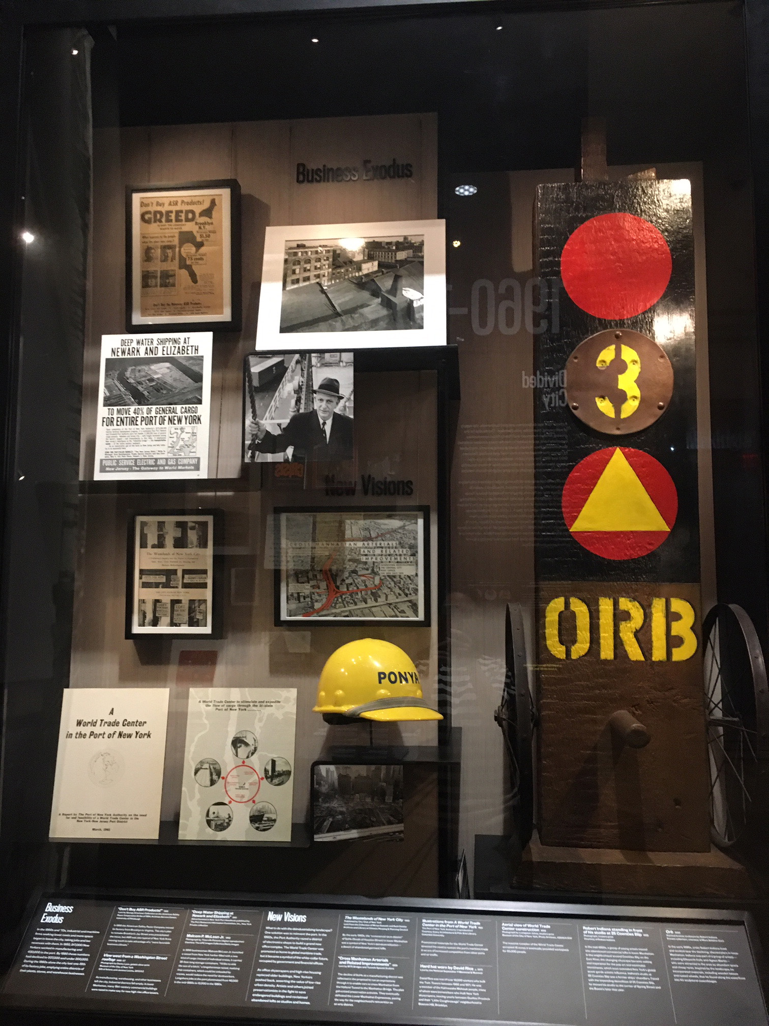 Museum display case with objects including a hard hat, framed photographs, and signage. 