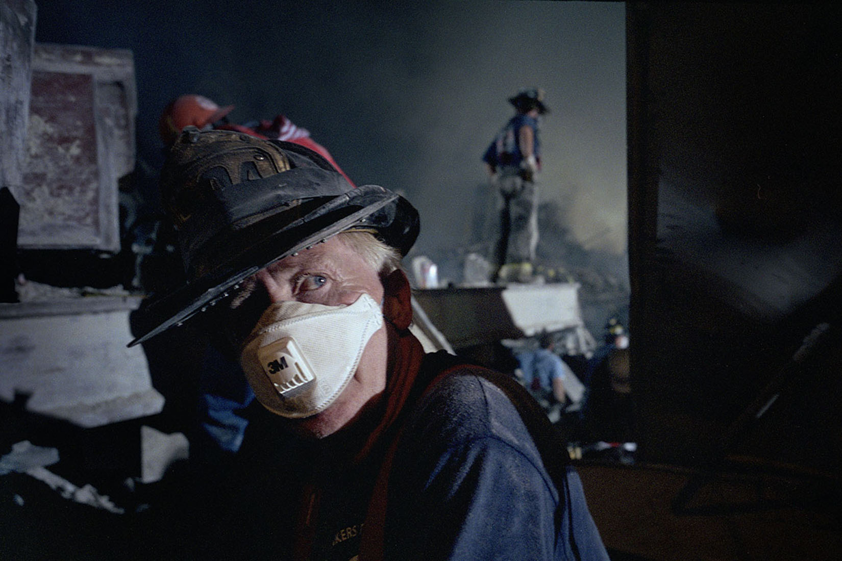 Closeup of a firefighter at Ground Zero in a soot-darkened helmet, wearing a respirator mask. 