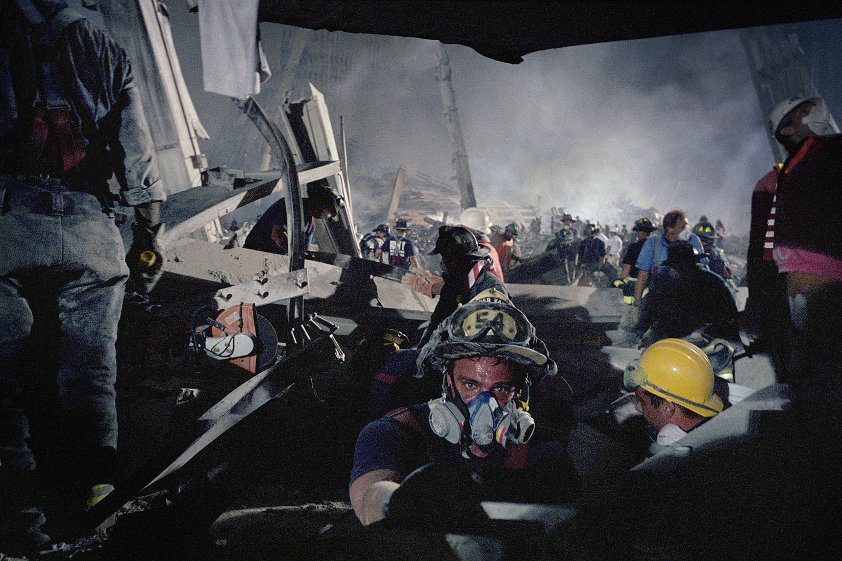 Photo of a firefighter, helmet number 54, with a sweaty forehead above a respiration mask, looking at the viewer from a pile of rubble. 