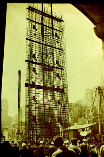 Longshot image of indiscernible people facing the vertically centered multi-story reinforcing steel cage.