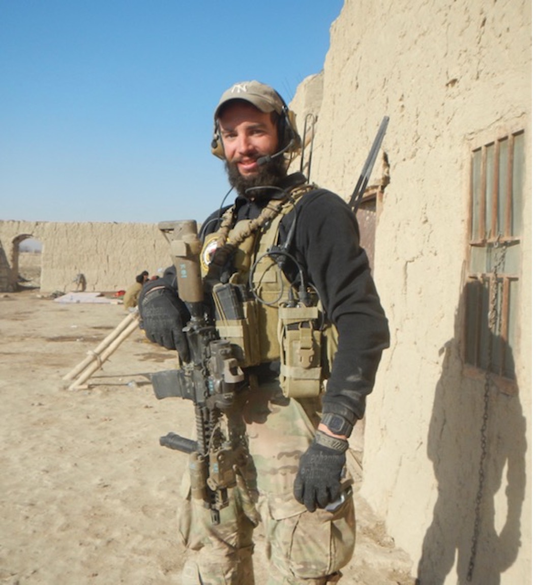 A bearded young man in an army uniform stands with weapons against a desert backdrop