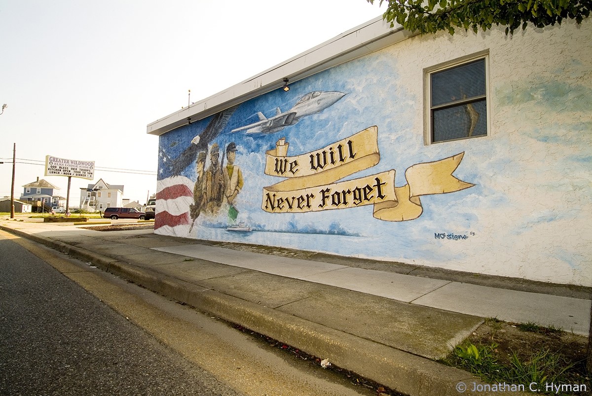 Mural featuring World War II-era ship on water; airplane above; servicemen; and a banner reading 'We Will Never Forget."