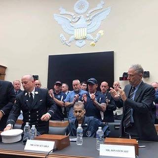 A man is seated at a table while two others stand to his left and right. Behind him are several rows of clapping people, in front of a row with the symbol of Congress.