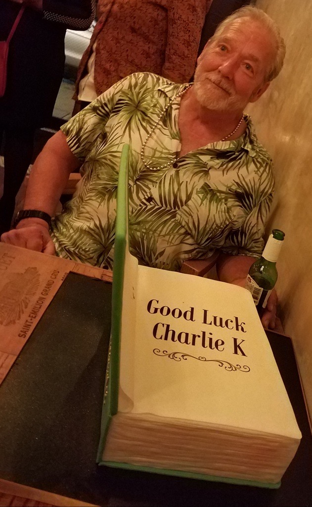 A smiling gray-haired man in a green and white Hawaiian print shirt behind a cake shaped like a book, with frosting that says Good Luck Charlie K