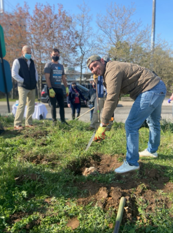 At right, a smiling male in blue jeans, a beige jacket, white sneakers, and a backwards baseball cap bends over to tend to small dirt hole in a patch of grass while others look on in the background