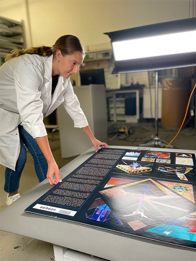 A woman in a ponytail, jeans, and white lab coat leans over to adjust boards depicting proposed memorial designs 