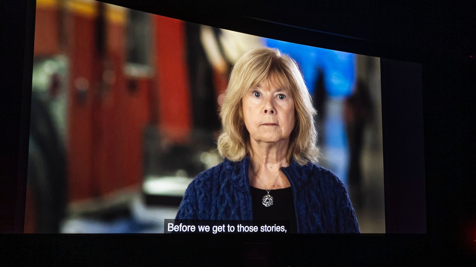 A woman with shoulder-length blonde hair appears on a movie screen, speaking to the audience. A subtitle is visible underneath her, and the surrounding theater is darkened.