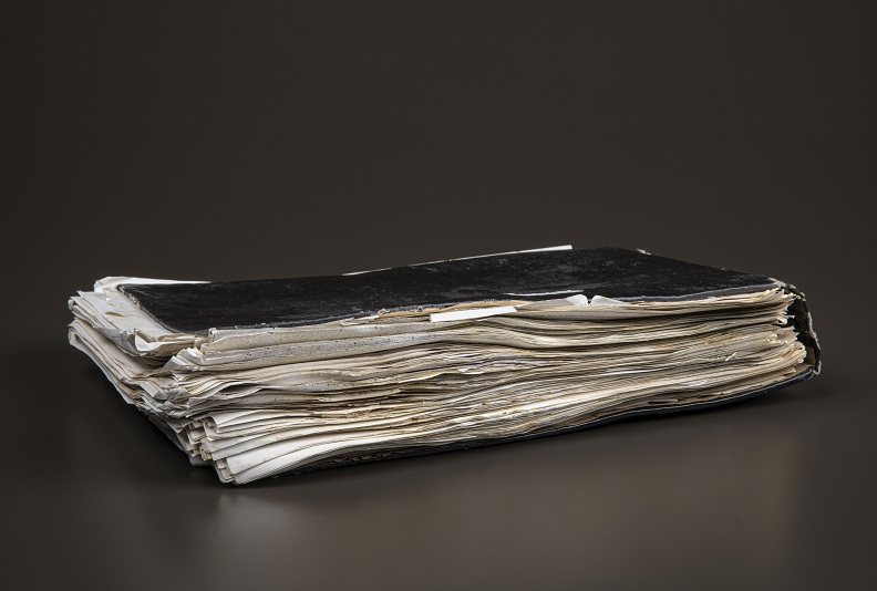 Binder with black cover and full with white documents slightly larger than the cover. It shows signs of wear with some tears.